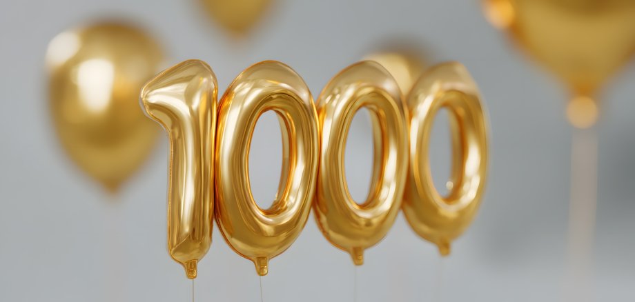Goldene Luftballons in Form der Zahl „1000“ schweben vor einem neutralen Hintergrund. Im unscharfen Hintergrund sind weitere goldene Ballons zu sehen – das Bild vermittelt festliche Stimmung und Jubiläumsfreude.