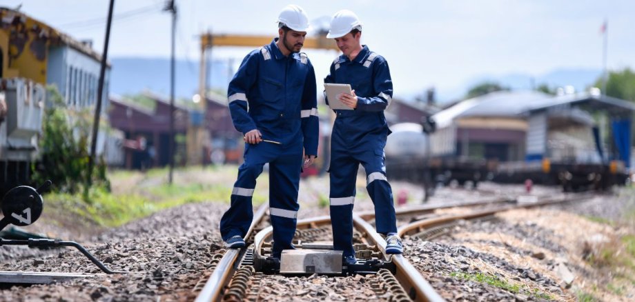 Zwei Arbeiter in blauer Schutzkleidung und weißen Helmen stehen auf Bahngleisen. Sie prüfen technische Daten auf einem Tablet. Im Hintergrund sind Züge, Werkstätten und Strommasten zu sehen.