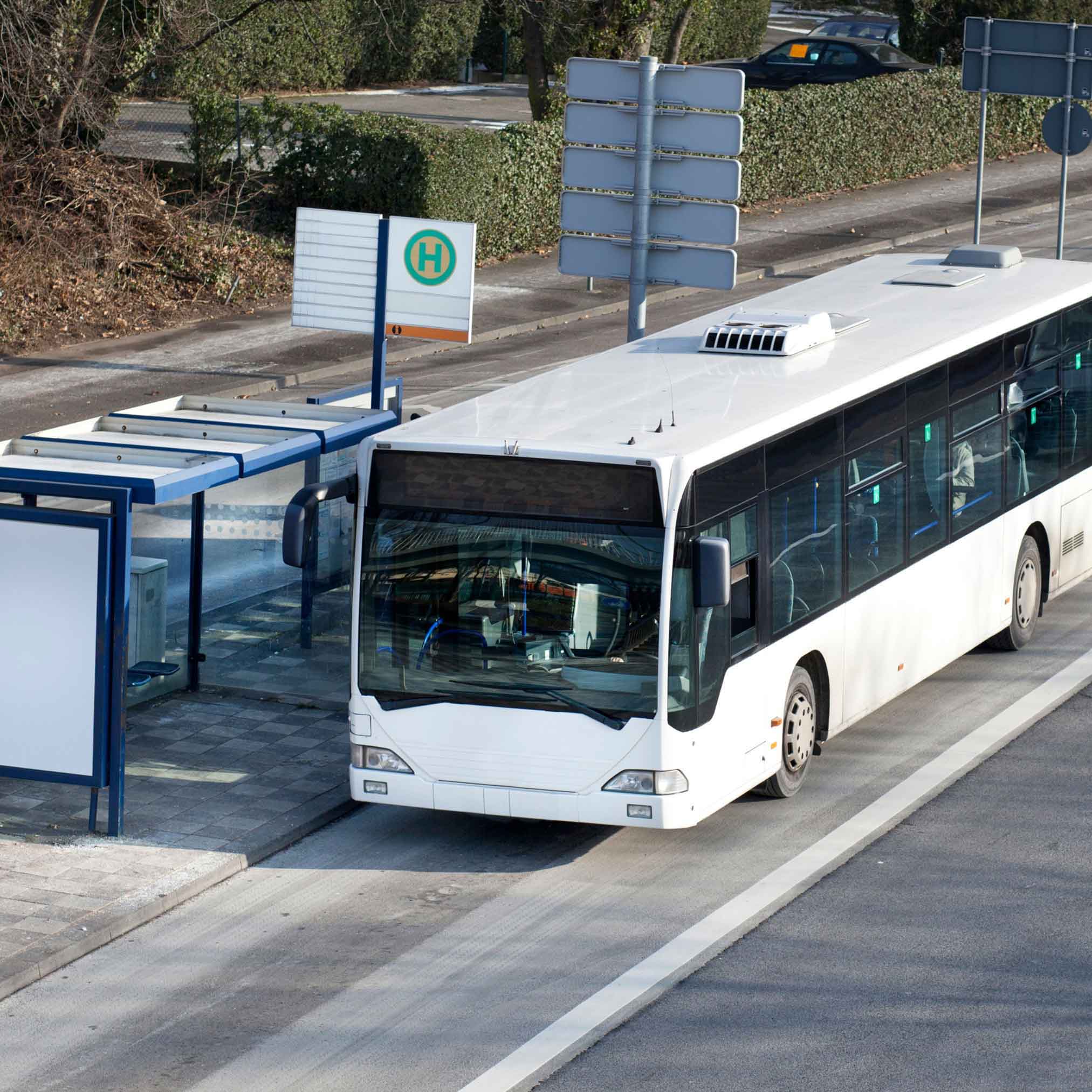 Ein weißer Linienbus hält an einer modernen Bushaltestelle mit überdachtem Wartebereich. Im Hintergrund sind Straßenschilder, Hecken und parkende Autos zu sehen. Das Bild wirkt ruhig und alltagsnah.