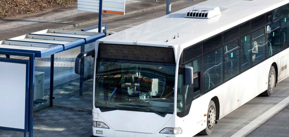 Ein weißer Linienbus hält an einer modernen Bushaltestelle mit überdachtem Wartebereich. Im Hintergrund sind Straßenschilder, Hecken und parkende Autos zu sehen. Das Bild wirkt ruhig und alltagsnah.