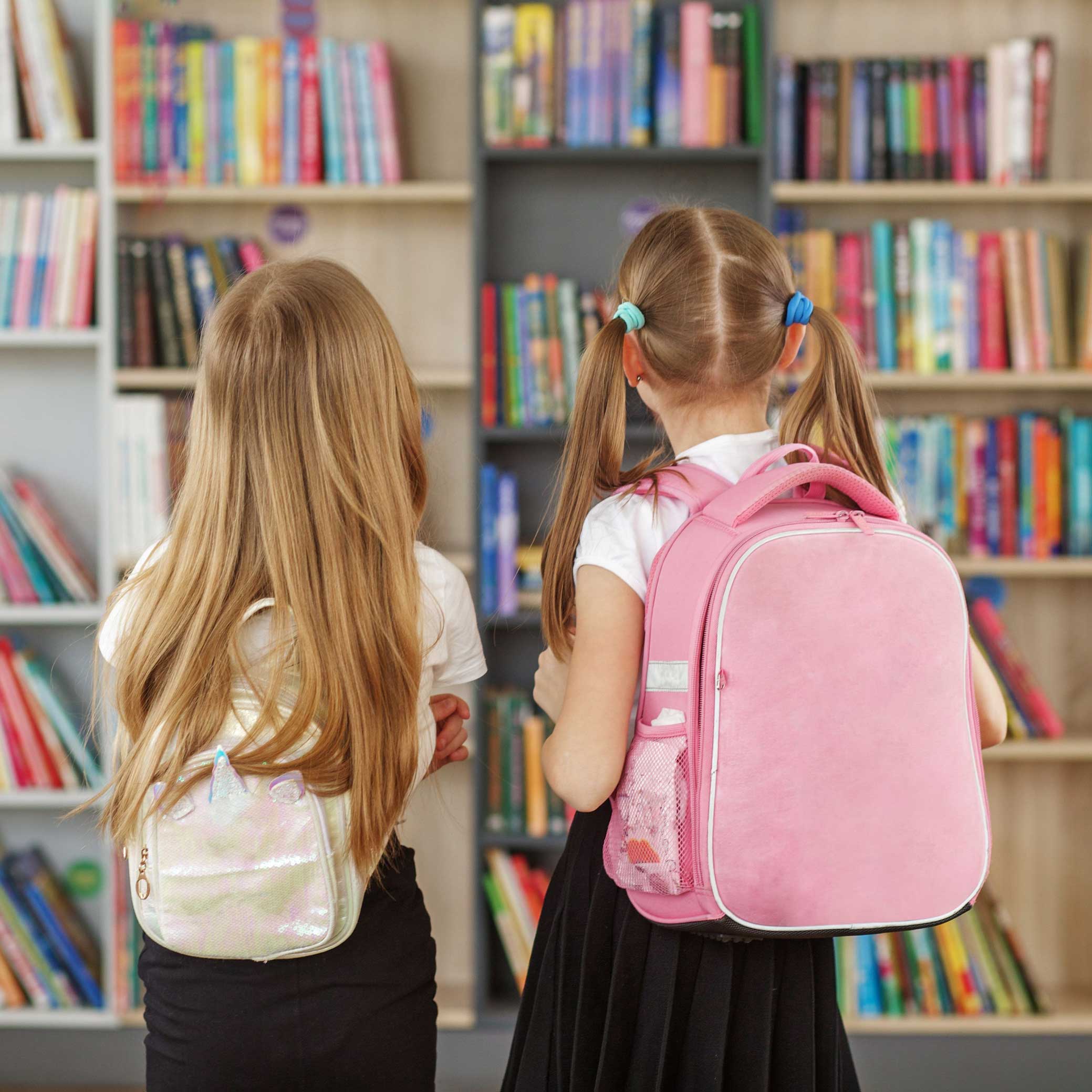 Zwei junge Mädchen mit Schulranzen stehen in einer Bücherei und betrachten bunte Bücherregale. Die Szene wirkt ruhig und neugierig – ein Moment kindlicher Entdeckerfreude beim Stöbern.