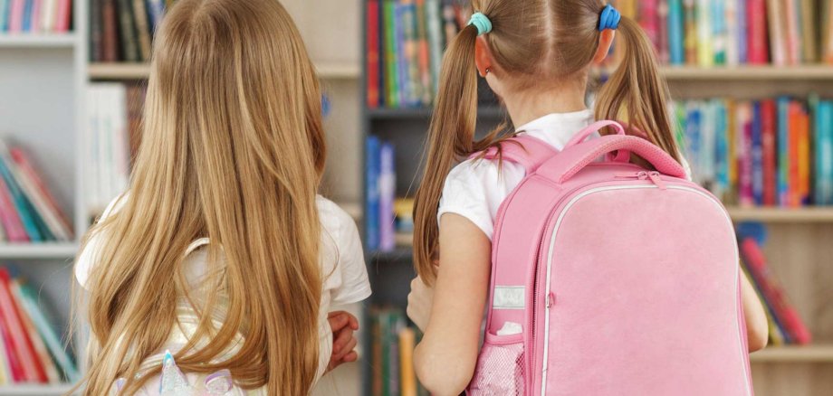 Zwei junge Mädchen mit Schulranzen stehen in einer Bücherei und betrachten bunte Bücherregale. Die Szene wirkt ruhig und neugierig – ein Moment kindlicher Entdeckerfreude beim Stöbern.