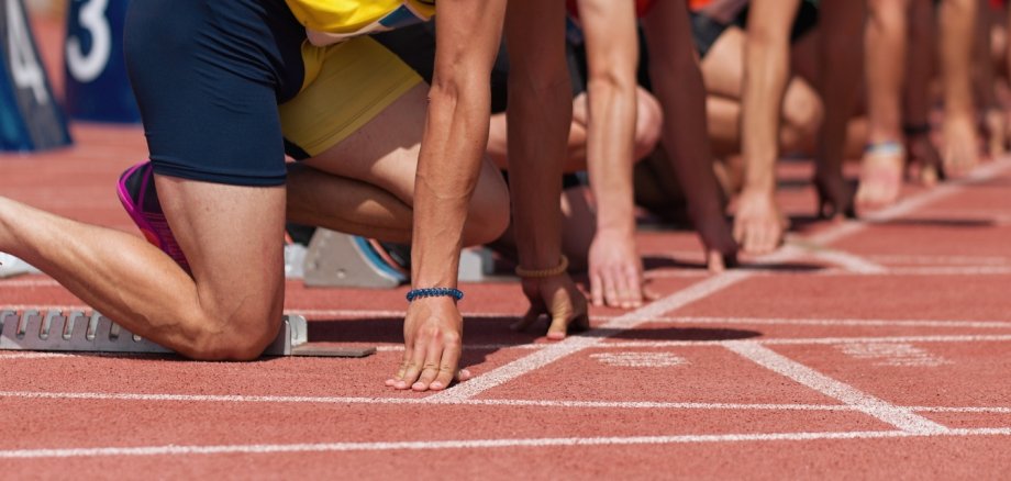Mehrere Läufer stehen in Startposition auf einer roten Tartanbahn. Der Fokus liegt auf den Beinen und Startblöcken. Gleich beginnt ein Wettlauf – Spannung und Konzentration liegen in der Luft.
