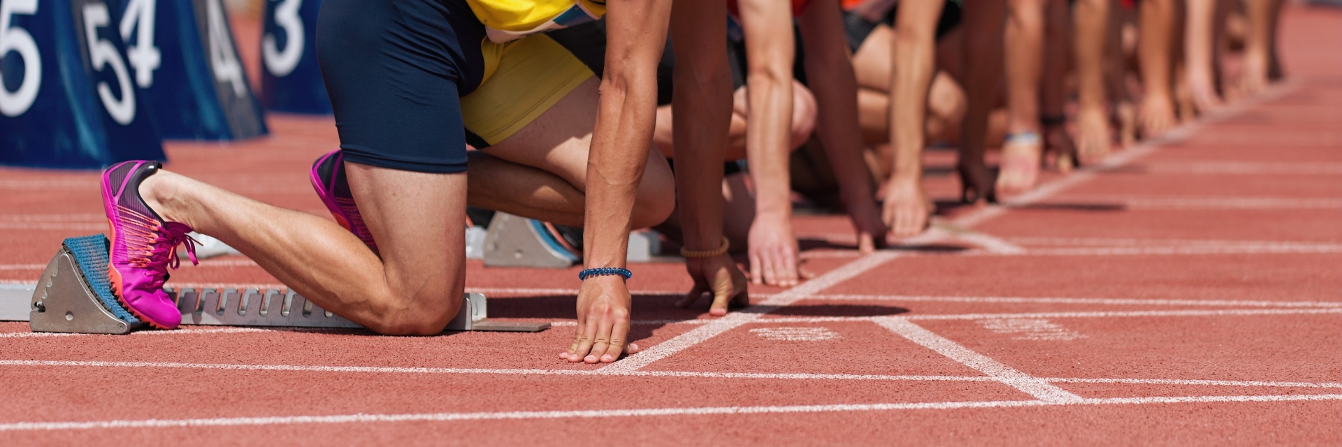 Mehrere Läufer stehen in Startposition auf einer roten Tartanbahn. Der Fokus liegt auf den Beinen und Startblöcken. Gleich beginnt ein Wettlauf – Spannung und Konzentration liegen in der Luft.
