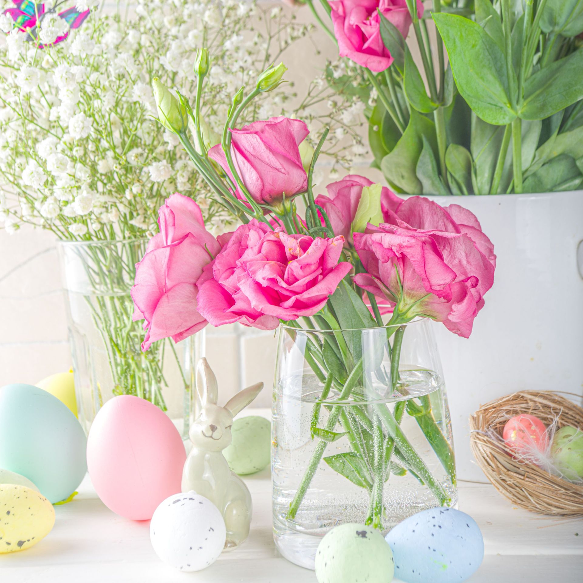 Ein frühlingshaft dekorierter Tisch mit rosa Blumen in einer Vase, bunten Ostereiern, einem kleinen Hasenfigur und einem Nest. Das Bild vermittelt eine helle, freundliche Osterstimmung.