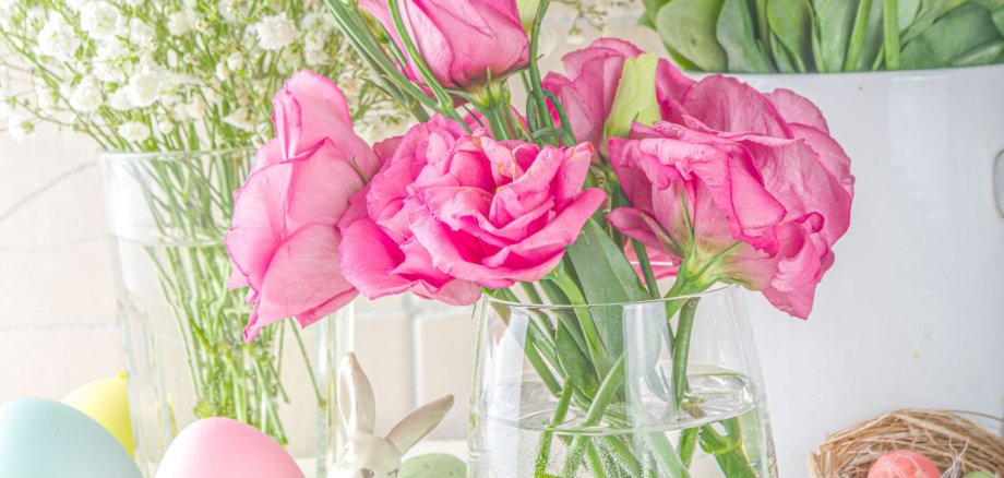 Ein frühlingshaft dekorierter Tisch mit rosa Blumen in einer Vase, bunten Ostereiern, einem kleinen Hasenfigur und einem Nest. Das Bild vermittelt eine helle, freundliche Osterstimmung.