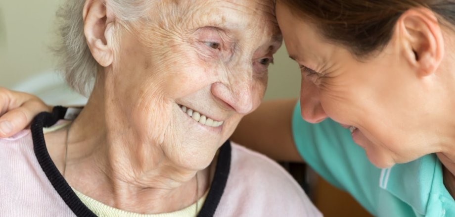 Eine Pflegerin und eine ältere Frau lächeln sich liebevoll an und berühren ihre Stirnen. Das Bild vermittelt Nähe, Vertrauen und Herzlichkeit – ein Moment menschlicher Zuwendung in der Pflege.