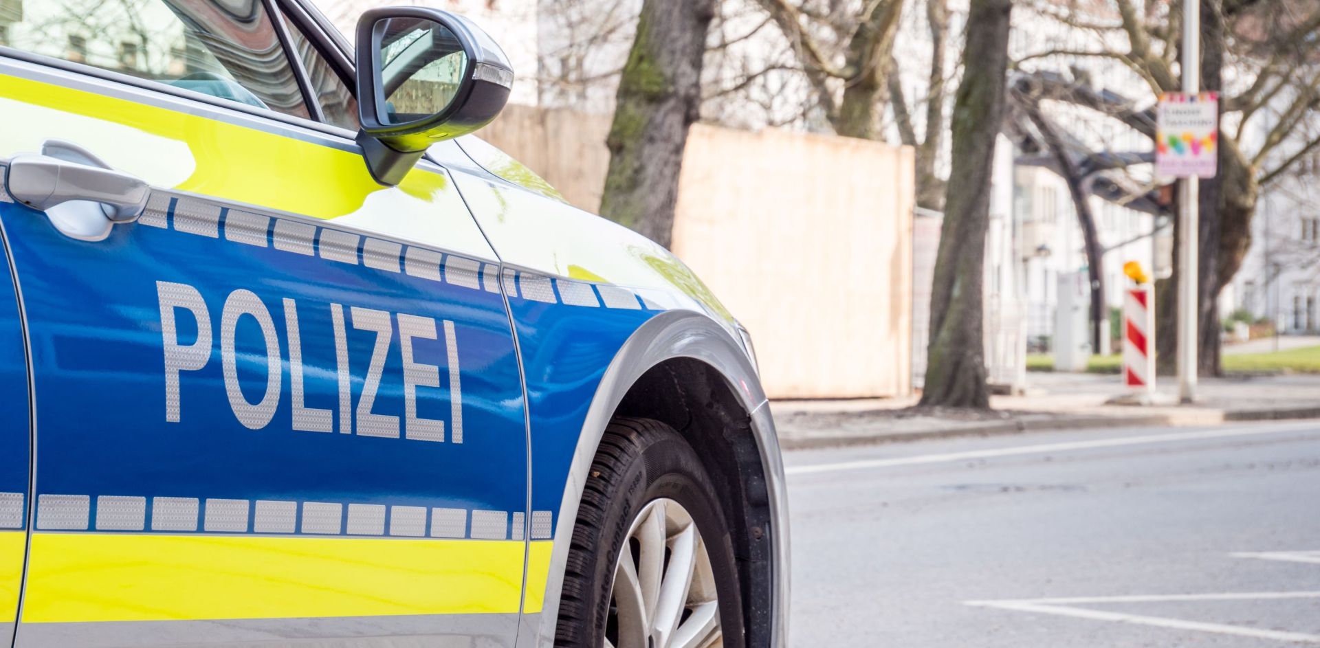 Ein blau-gelbes Polizeiauto steht am Straßenrand in einer städtischen Umgebung. Auf der Fahrzeugseite ist deutlich das Wort „Polizei“ zu lesen. Im Hintergrund sind Bäume und Gebäude zu sehen.