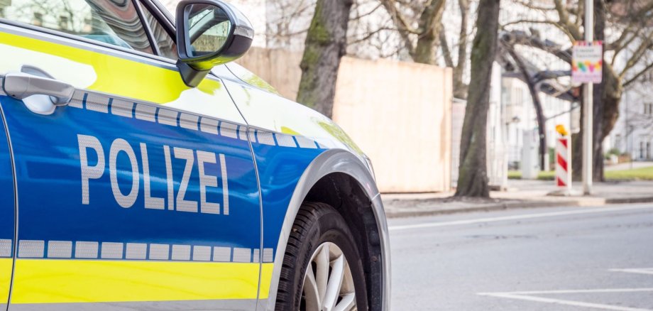 Ein blau-gelbes Polizeiauto steht am Straßenrand in einer städtischen Umgebung. Auf der Fahrzeugseite ist deutlich das Wort „Polizei“ zu lesen. Im Hintergrund sind Bäume und Gebäude zu sehen.
