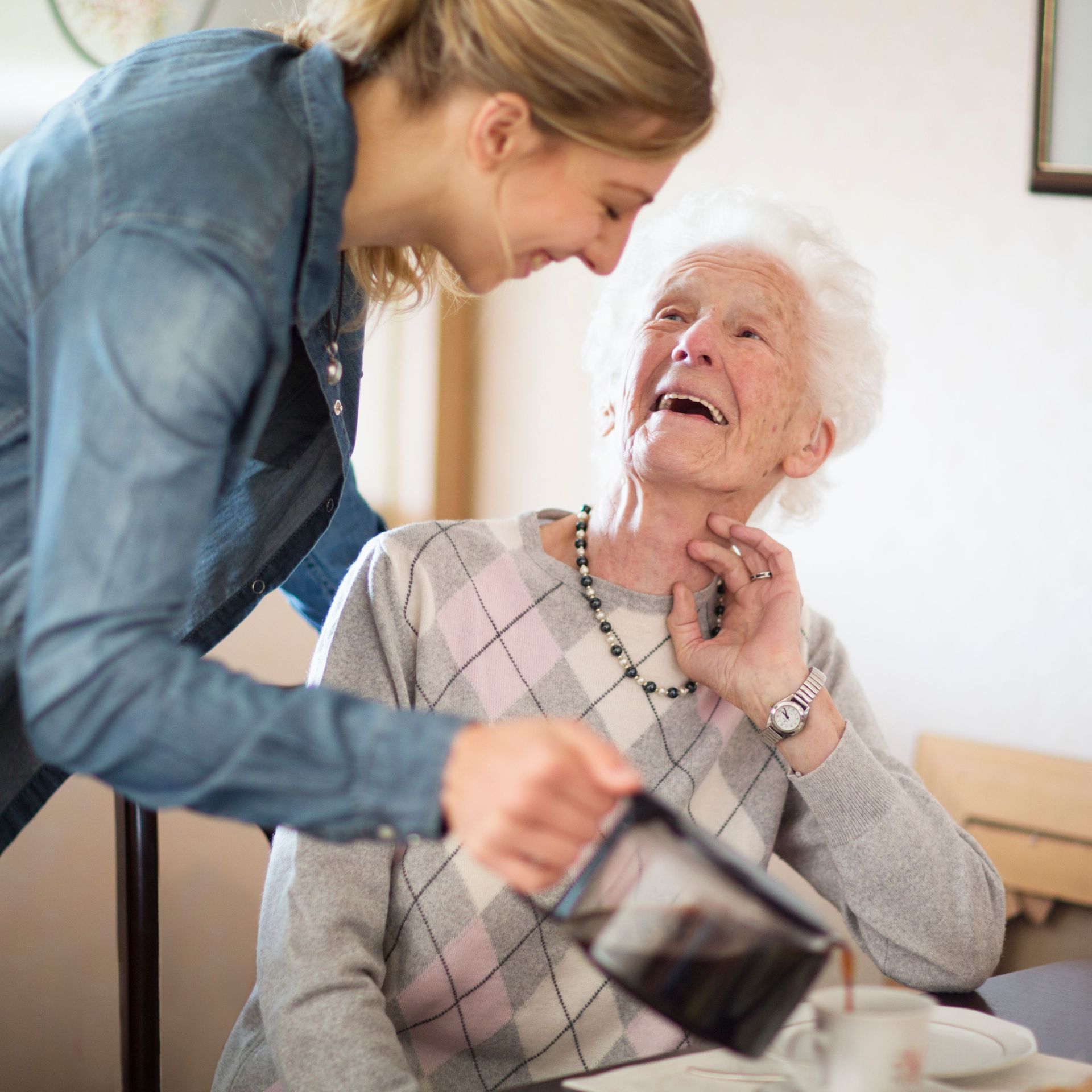 Eine junge Frau schenkt einer älteren Dame lächelnd Kaffee ein. Beide wirken fröhlich und herzlich verbunden. Das Bild strahlt Wärme, Fürsorge und zwischenmenschliche Nähe im Alltag aus.
