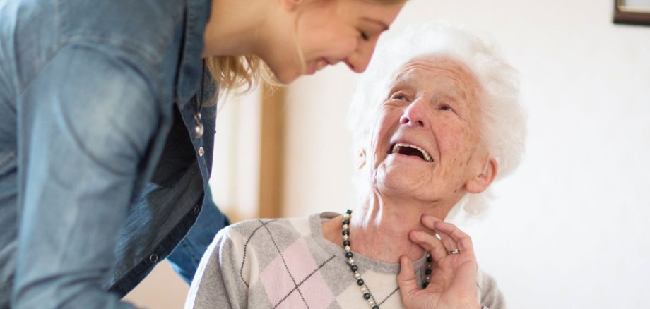 Eine junge Frau schenkt einer älteren Dame lächelnd Kaffee ein. Beide wirken fröhlich und herzlich verbunden. Das Bild strahlt Wärme, Fürsorge und zwischenmenschliche Nähe im Alltag aus.