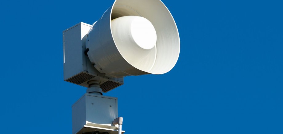 Eine moderne Sirene ist auf einem Mast montiert und ragt vor einem klaren blauen Himmel empor. Das Bild symbolisiert Katastrophenschutz, Warnsysteme und die Bedeutung öffentlicher Sicherheitsinfrastruktur.