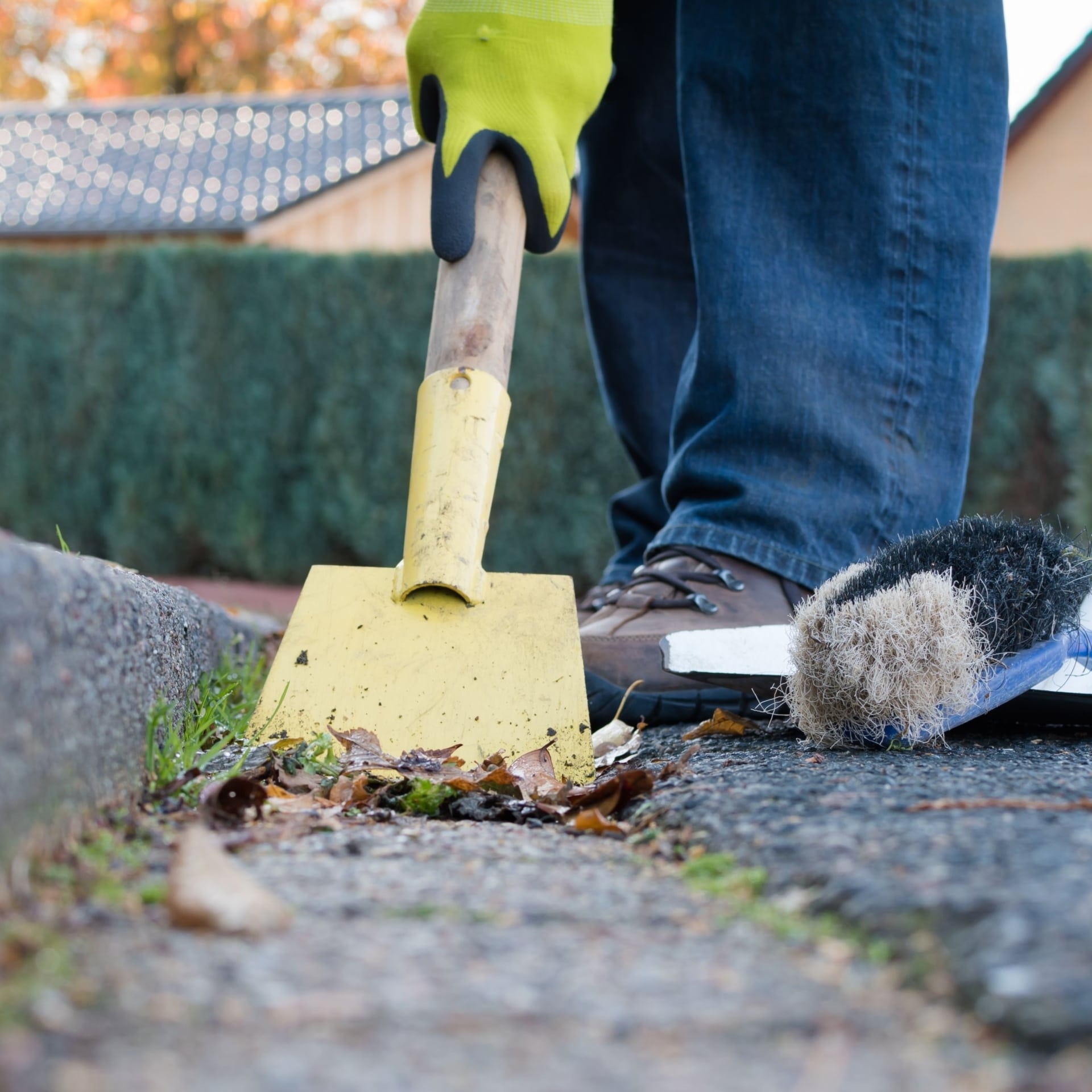 Eine Person mit Arbeitshandschuhen und robusten Schuhen reinigt mit einer gelben Schaufel und einer Bürste den Straßenrand. Auf dem Boden liegen Blätter und Schmutz – herbstliche Straßenreinigung.