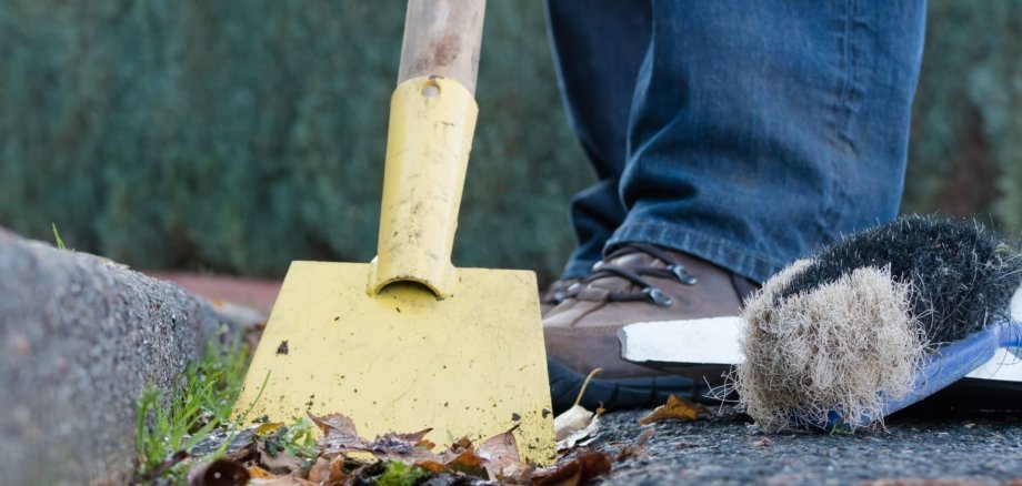 Eine Person mit Arbeitshandschuhen und robusten Schuhen reinigt mit einer gelben Schaufel und einer Bürste den Straßenrand. Auf dem Boden liegen Blätter und Schmutz – herbstliche Straßenreinigung.