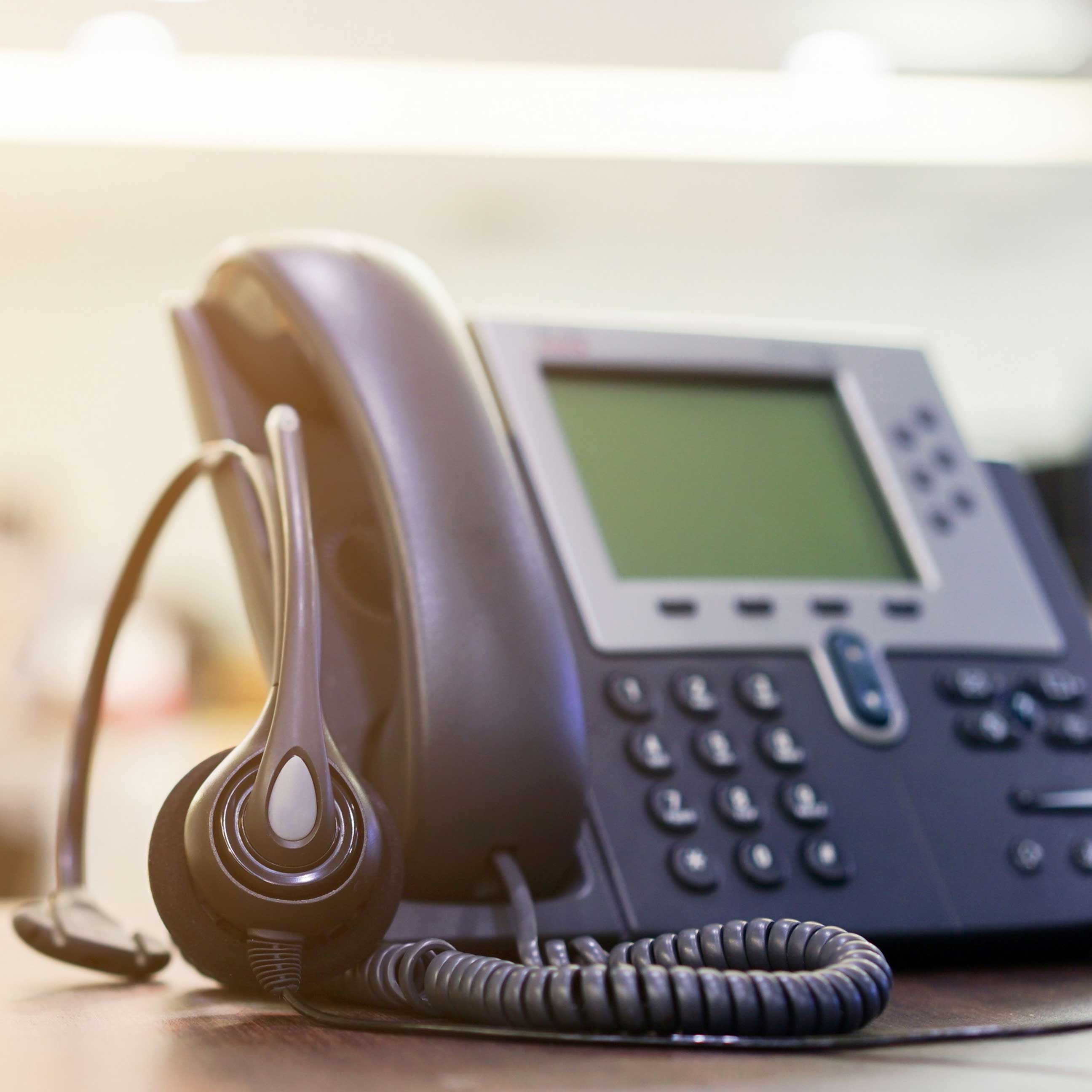 Ein modernes Bürotelefon mit Display steht auf einem Schreibtisch. Daneben liegt ein Headset mit Mikrofon. Die Szene wirkt professionell und symbolisiert Kommunikation, Service oder telefonische Erreichbarkeit.