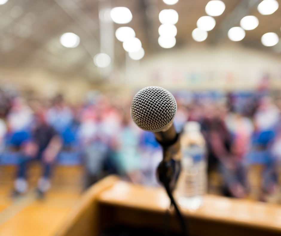Ein Mikrofon steht auf einem Rednerpult, dahinter ist eine unscharfe Menschenmenge in einer Halle zu sehen. Das Bild vermittelt die Atmosphäre einer Versammlung, Rede oder öffentlichen Veranstaltung.