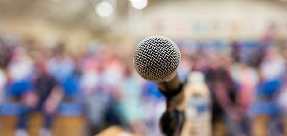 Ein Mikrofon steht auf einem Rednerpult, dahinter ist eine unscharfe Menschenmenge in einer Halle zu sehen. Das Bild vermittelt die Atmosphäre einer Versammlung, Rede oder öffentlichen Veranstaltung.