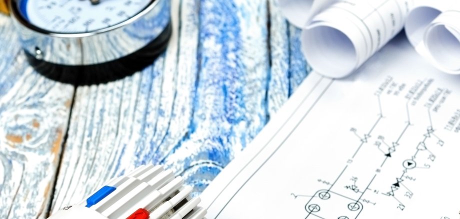 Auf einem Holztisch liegen technische Pläne, ein Thermostat, Messinstrumente und Heizungsbauteile aus Metall. Die Szene symbolisiert Wärmeplanung, Energieeffizienz und moderne Gebäudetechnik im Bereich Heizung und Klimasysteme.