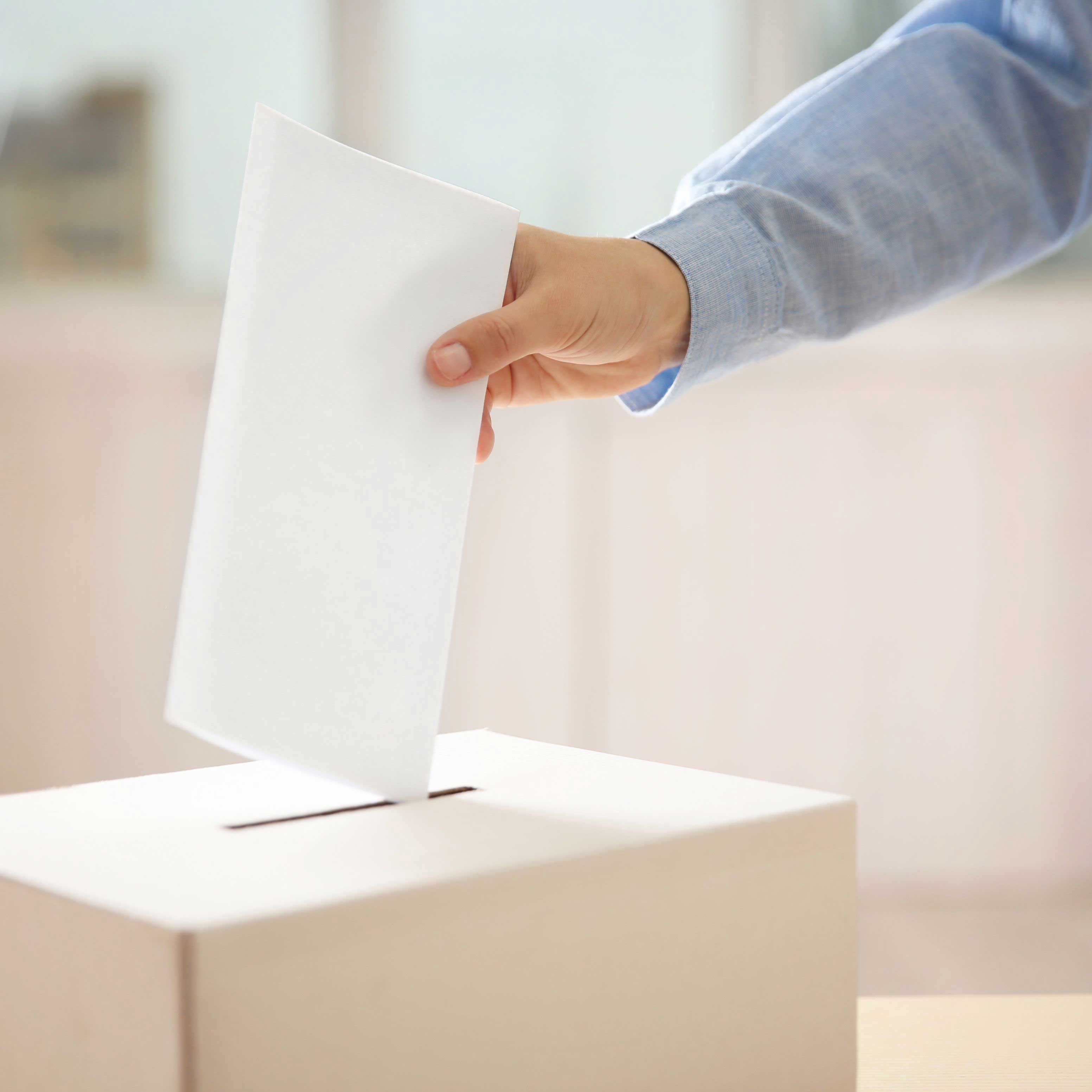 Eine Person steckt einen weißen Umschlag in eine Wahlurne. Die Szene symbolisiert eine Stimmabgabe bei einer Wahl. Der Hintergrund ist hell und neutral, der Fokus liegt auf der Handbewegung.