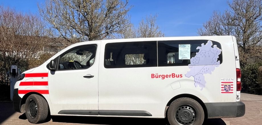 Ein weißer Kleinbus mit der Aufschrift „BürgerBus“ steht auf einem gepflasterten Platz. Auf der Seite sind das Hessen-Wappen und eine Karte des Bundeslandes zu sehen. Blauer Himmel, Sonnenschein.