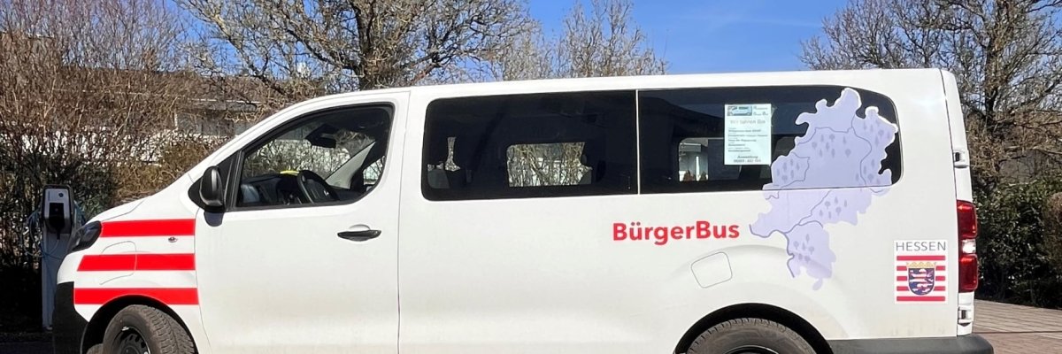 Ein weißer Kleinbus mit der Aufschrift „BürgerBus“ steht auf einem gepflasterten Platz. Auf der Seite sind das Hessen-Wappen und eine Karte des Bundeslandes zu sehen. Blauer Himmel, Sonnenschein.