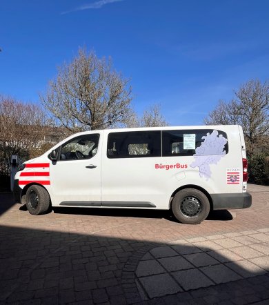 Ein weißer Kleinbus mit der Aufschrift „BürgerBus“ steht auf einem gepflasterten Platz. Auf der Seite sind das Hessen-Wappen und eine Karte des Bundeslandes zu sehen. Blauer Himmel, Sonnenschein.