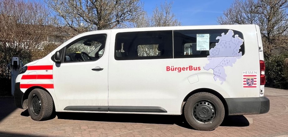 Ein weißer Kleinbus mit der Aufschrift „BürgerBus“ steht auf einem gepflasterten Platz. Auf der Seite sind das Hessen-Wappen und eine Karte des Bundeslandes zu sehen. Blauer Himmel, Sonnenschein.