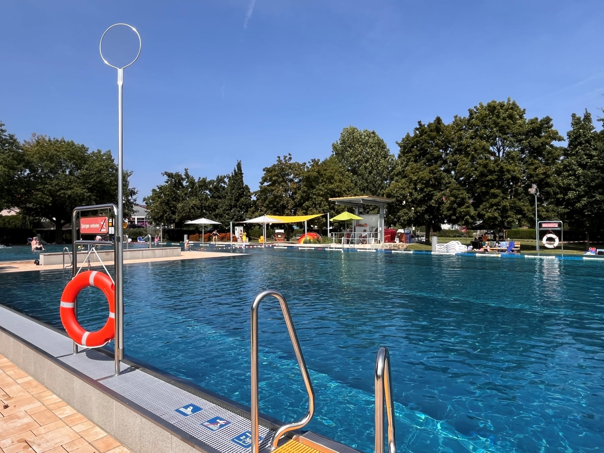 Das Foto zeigt das Freibad Rodheim an einem sonnigen Tag. Klare Wasseroberfläche, Rettungsringe und Sonnenschirme prägen die Szene. Besucher entspannen am Beckenrand, umgeben von grünen Bäumen und blauem Himmel.