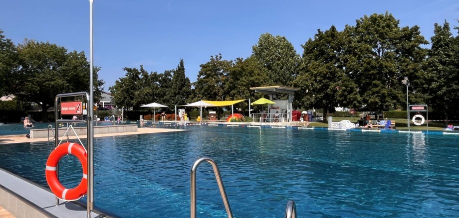 Das Foto zeigt das Freibad Rodheim an einem sonnigen Tag. Klare Wasseroberfläche, Rettungsringe und Sonnenschirme prägen die Szene. Besucher entspannen am Beckenrand, umgeben von grünen Bäumen und blauem Himmel.