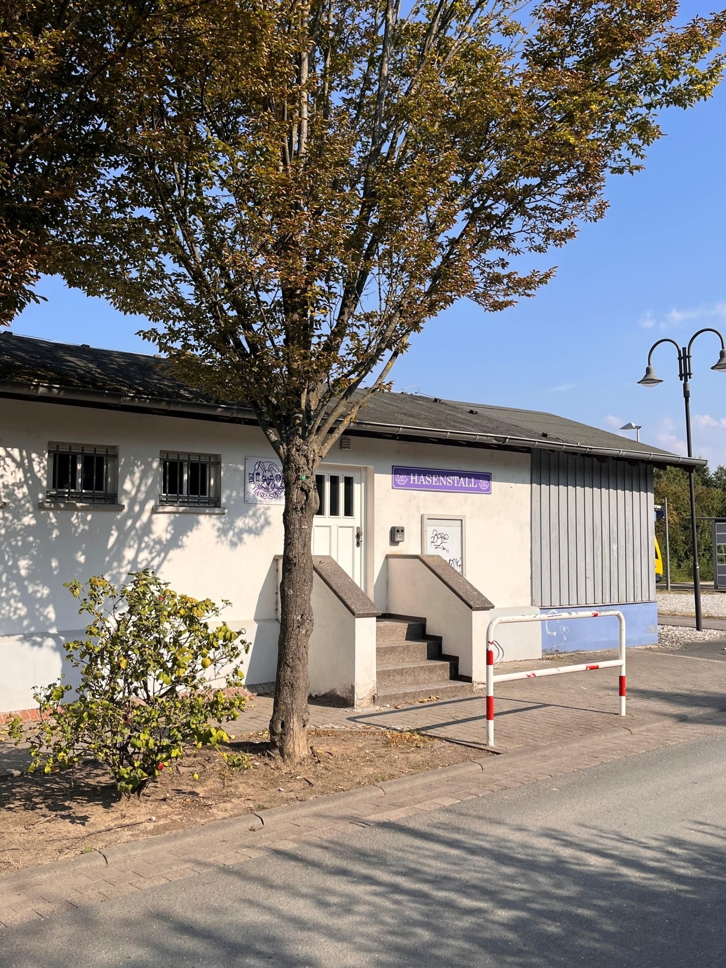 Das Foto zeigt ein helles Gebäude mit der Aufschrift „Hasenstall“ in lila Schrift. Vor dem Eingang stehen ein Baum, eine Treppe und eine rot-weiße Absperrung. Sommerliche Atmosphäre.