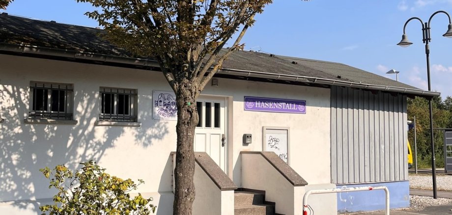 Das Foto zeigt ein helles Gebäude mit der Aufschrift „Hasenstall“ in lila Schrift. Vor dem Eingang stehen ein Baum, eine Treppe und eine rot-weiße Absperrung. Sommerliche Atmosphäre.