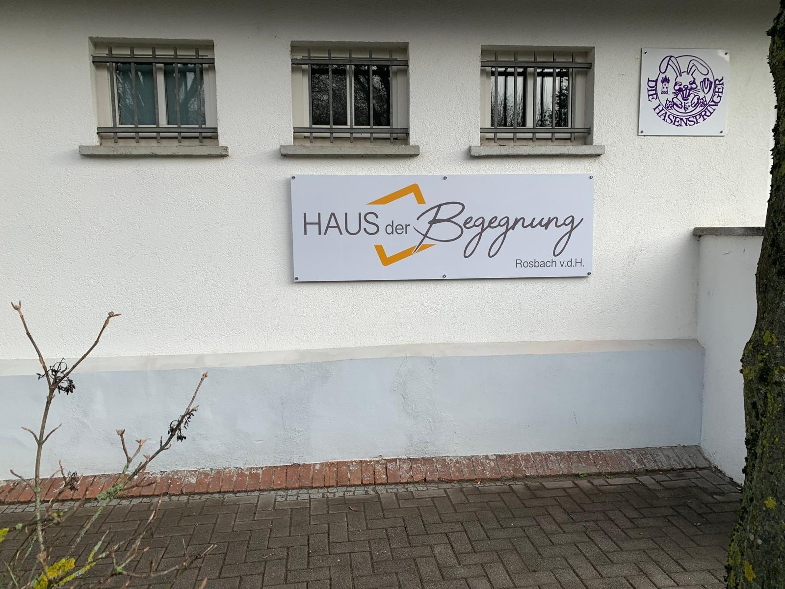 An einer weißen Hauswand hängt ein Schild mit der Aufschrift „Haus der Begegnung Rosbach v. d. H.“. Daneben ist das Logo des Vereins „Die Hasenspringer“ zu sehen.