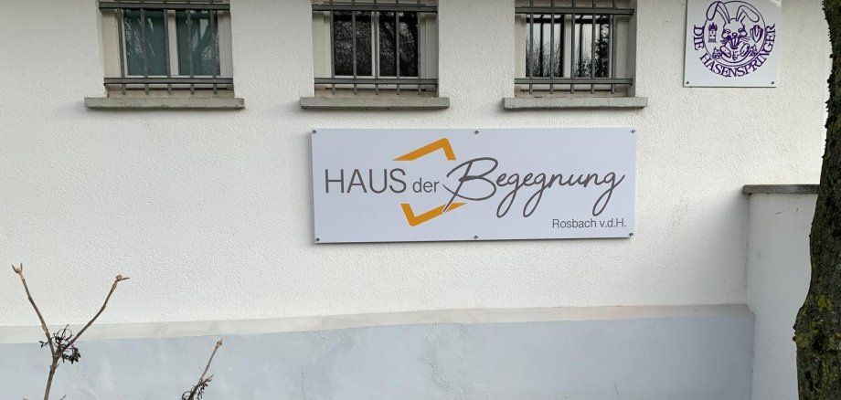 An einer weißen Hauswand hängt ein Schild mit der Aufschrift „Haus der Begegnung Rosbach v. d. H.“. Daneben ist das Logo des Vereins „Die Hasenspringer“ zu sehen.