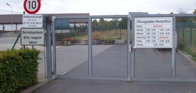 Das Bild zeigt das verschlossene Tor des Recyclinghofs. An der Einfahrt hängen Schilder mit Öffnungszeiten, Geschwindigkeitsbegrenzung und Hinweisen zum Betriebsgelände. Dahinter sind Container und Holzstapel zu sehen.