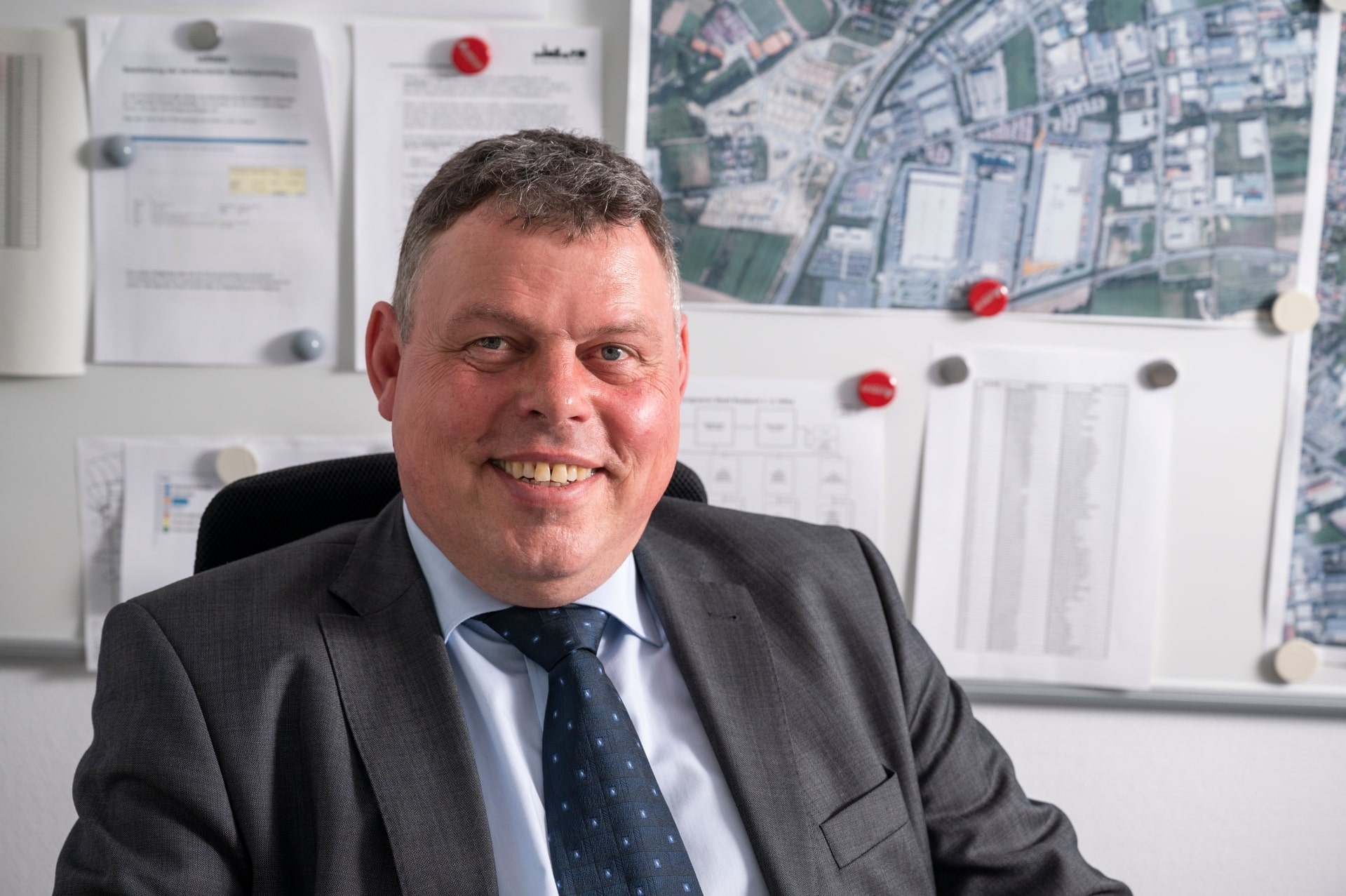 Bürgermeister Steffen Maar sitzt im Anzug sitzt lächelnd in seinem Büro vor einer Pinnwand mit Stadtplänen und Dokumenten. Das Bild vermittelt einen professionellen, freundlichen Eindruck in einer kommunalen Arbeitsumgebung.