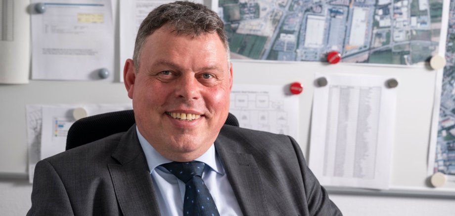 Bürgermeister Steffen Maar sitzt im Anzug sitzt lächelnd in seinem Büro vor einer Pinnwand mit Stadtplänen und Dokumenten. Das Bild vermittelt einen professionellen, freundlichen Eindruck in einer kommunalen Arbeitsumgebung.