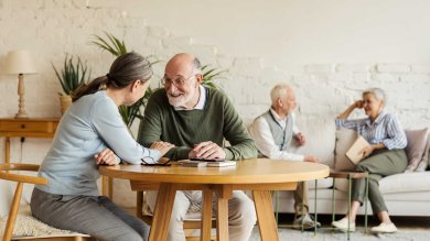Zwei ältere Personen sitzen an einem Holztisch und unterhalten sich lächelnd. Im unscharfen Hintergrund unterhalten sich weitere Senioren auf einer Couch vor einer hellen Ziegelwand, was eine soziale Atmosphäre vermittelt.