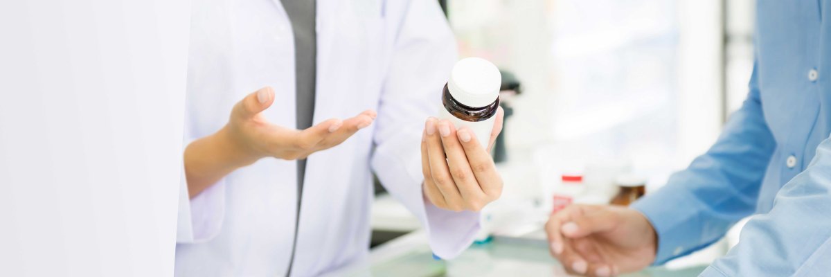 Eine Person in weißem Kittel (Apotheker/in) reicht einem Kunden mit blauem Hemd eine weiße Medikamentenflasche über den Tresen. Der Fokus liegt auf der Übergabe der Flasche.