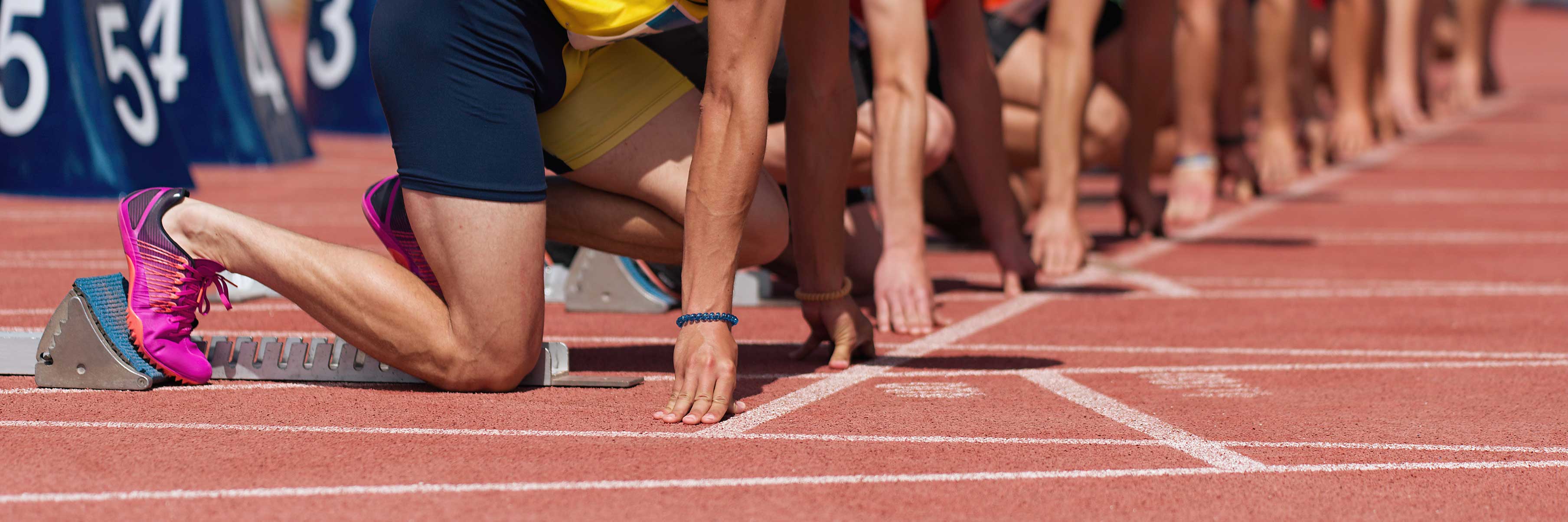 Mehrere Läufer in Startblöcken auf einer roten Leichtathletikbahn, bereit für den Start. Ihre Hände sind auf dem Boden, die Füße in den Blöcken. Startnummern sind im Hintergrund erkennbar.