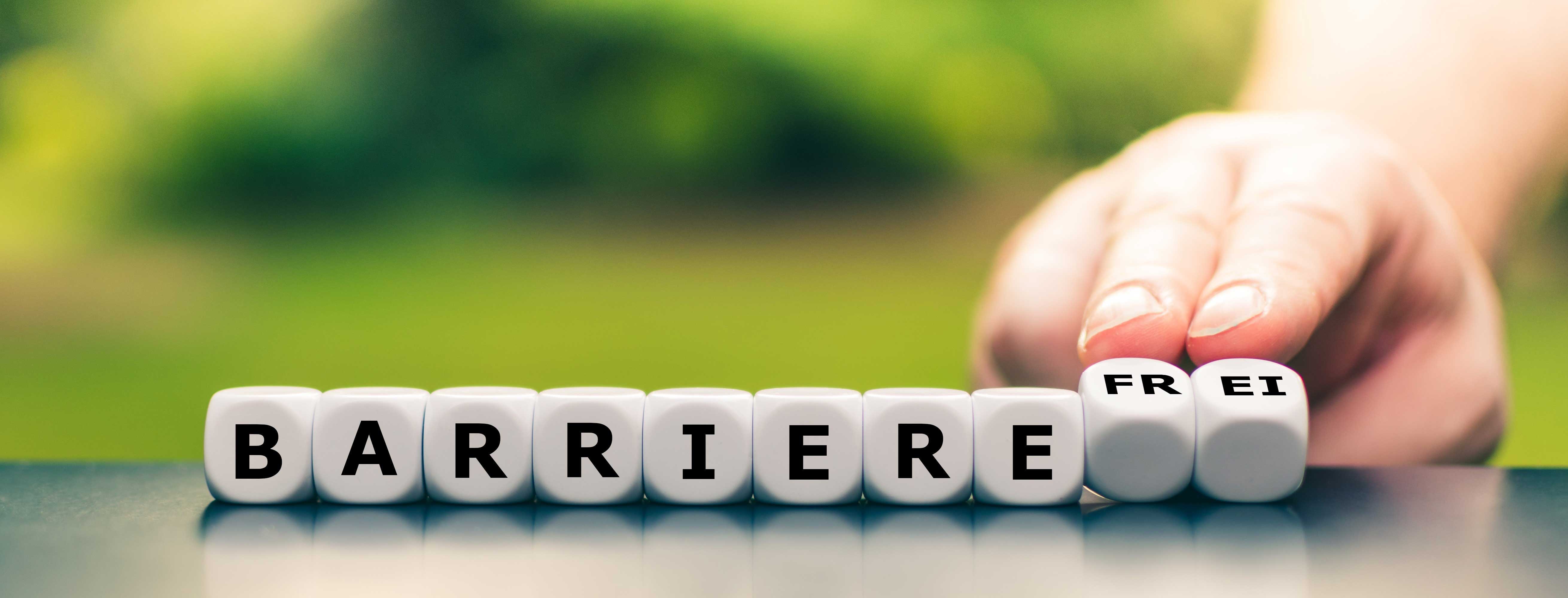 Weiße Würfel auf einem dunklen Untergrund buchstabieren "BARRIERE". Eine Hand schiebt die Würfel "FREI" hinzu, um das Wort "BARRIEREFREI" zu vervollständigen. Der Hintergrund ist unscharf grün.