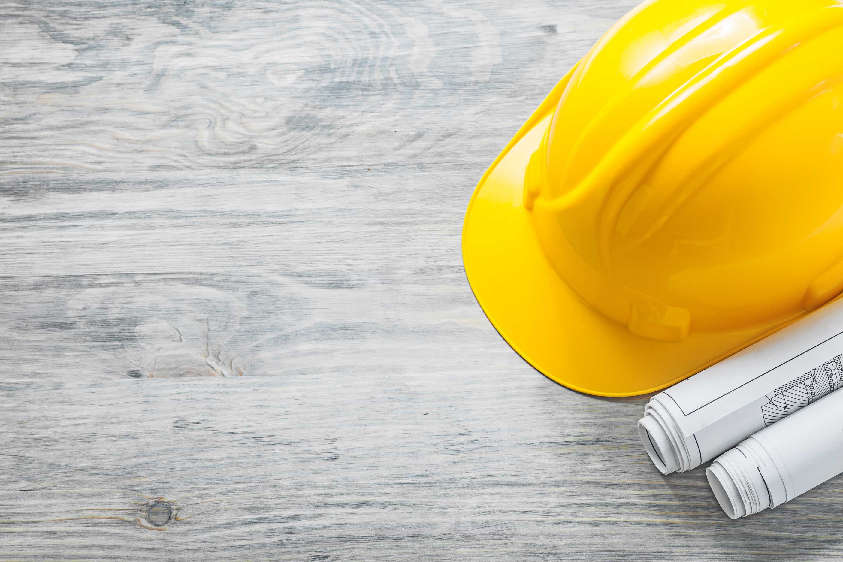 Ein gelber Bauhelm liegt neben gerollten Bauplänen auf einer hellen Holzoberfläche. Die Anordnung deutet auf Planung im Bauwesen hin.