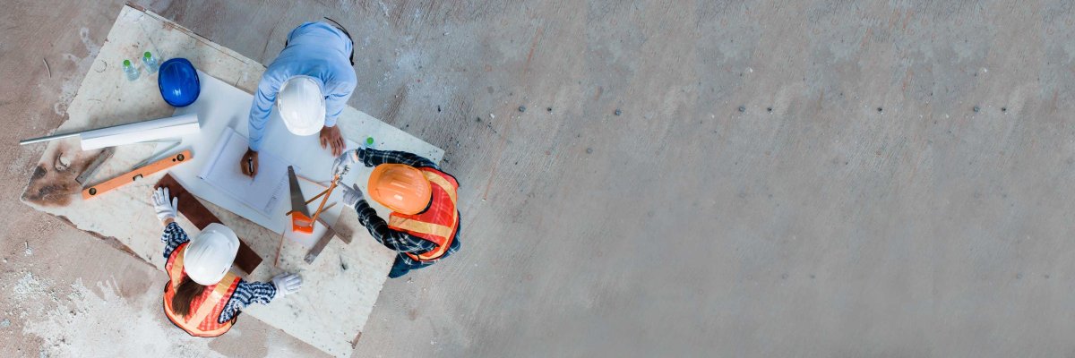 Drei Bauarbeiter in Schutzhelmen und Warnwesten betrachten von oben Baupläne auf einer Arbeitsplatte. Werkzeuge wie Säge und Wasserwaage liegen daneben.