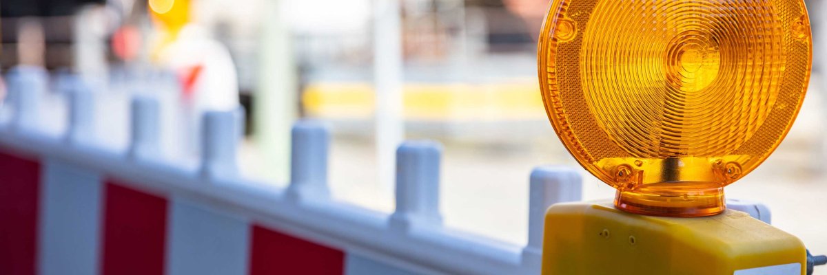 Auf einer rot-weiß gestreiften Baustellenabsperrung ist ein orangefarbenes Warnlicht befestigt. Im unscharfen Hintergrund ist eine typische Baustellenumgebung in der Stadt zu erkennen.