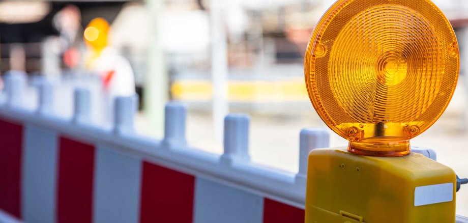 Auf einer rot-weiß gestreiften Baustellenabsperrung ist ein orangefarbenes Warnlicht befestigt. Im unscharfen Hintergrund ist eine typische Baustellenumgebung in der Stadt zu erkennen.