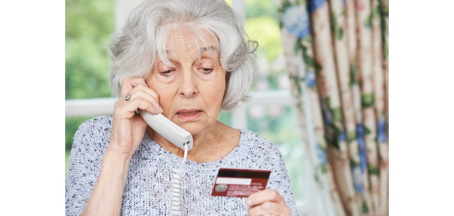 Eine ältere Frau telefoniert mit einem Festnetztelefon und hält mit besorgtem Ausdruck eine Bank- oder Kreditkarte in der Hand, was auf einen möglichen Betrugsversuch hindeutet.
