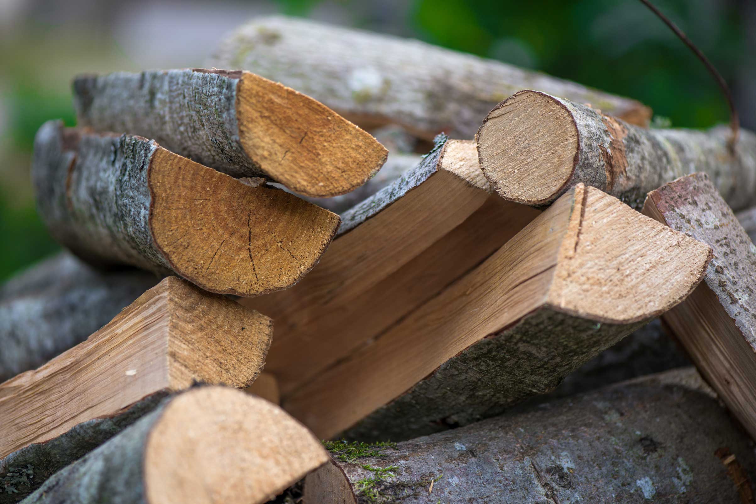 Ein detailreicher Holzstapel aus gespaltenen und ganzen Holzscheiten mit rissiger Rinde und sichtbaren Jahresringen. Der Hintergrund ist unscharf grün, was auf eine natürliche Umgebung hindeutet.