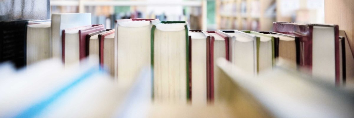Dicht aneinandergereihte Bücher mit hellen und roten Buchrücken stehen in einem Regal. Im unscharfen Hintergrund sind weitere Bücherregale einer Bibliothek zu erkennen.