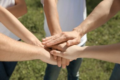 Mehrere Hände von verschiedenen Personen sind übereinandergelegt, die Fäuste nach unten gerichtet. Es symbolisiert Teamwork, Zusammenhalt und Verbundenheit, mit grünem Gras im unscharfen Hintergrund.