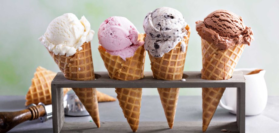 Vier Waffeltüten mit je einer Eiskugel (Vanille, Erdbeere, Stracciatella und Schokolade) stehen in einem Holzhalter auf einer Oberfläche mit bunten Streuseln.