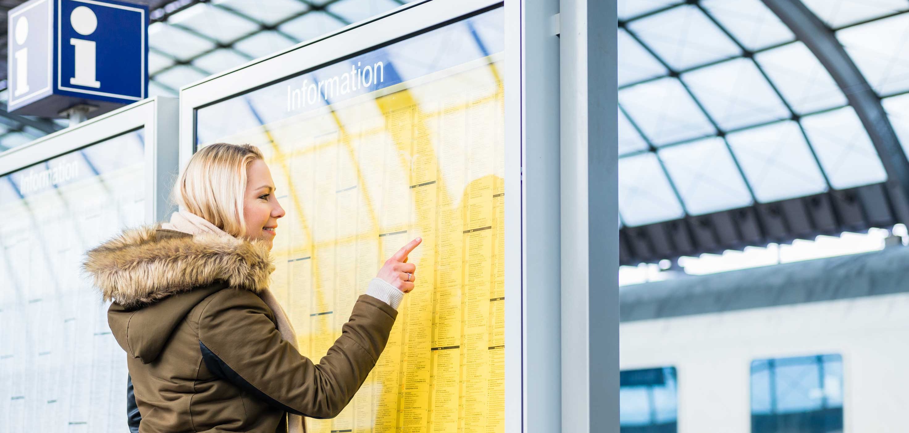 Eine blonde Frau in Winterkleidung steht an einem Bahnhof und zeigt auf eine gelbe Informationstafel. Im Hintergrund ist unscharf ein Zug und die Dachkonstruktion der Bahnhofshalle zu erkennen.